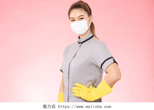 粉色背景上的清洁工女人迷人的亚洲女佣，戴着面罩，戴着手套，面带微笑，快乐而自信地准备着清洁，被粉色背景隔离，清洁服务的理念